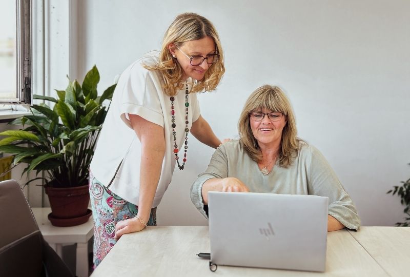 Zwei Frauen bei der Zusammenarbeit in einem Besprechungsraum. Eine Frau steht neben der sitzenden Frau, welche auf einen Laptop zeigt.