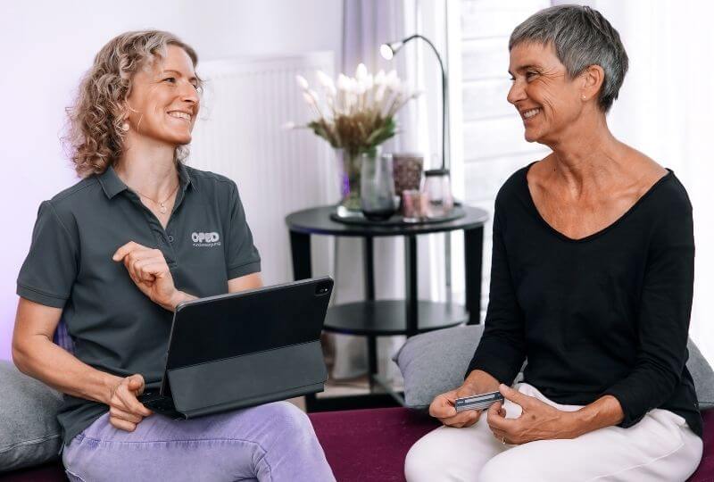 Bild stellt die OPED Wundversorgung als Teil der OPED Group dar. Zwei Frauen sitzend auf einer Couch. Eine von ihnen hat ein Tablet auf den Beinen und läüchelt der anderen Frau zu. Diese hat eine Krankenkarte in der Hand.